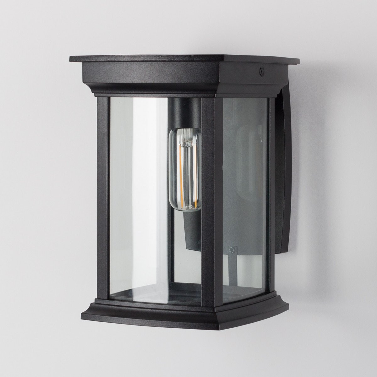 Zwarte wandlamp voor buiten, vierkante muursteun, lantaarnkap met zwart frame en heldere ruitjes, lichtbron boven in de kap, zichtbaar in het armatuur