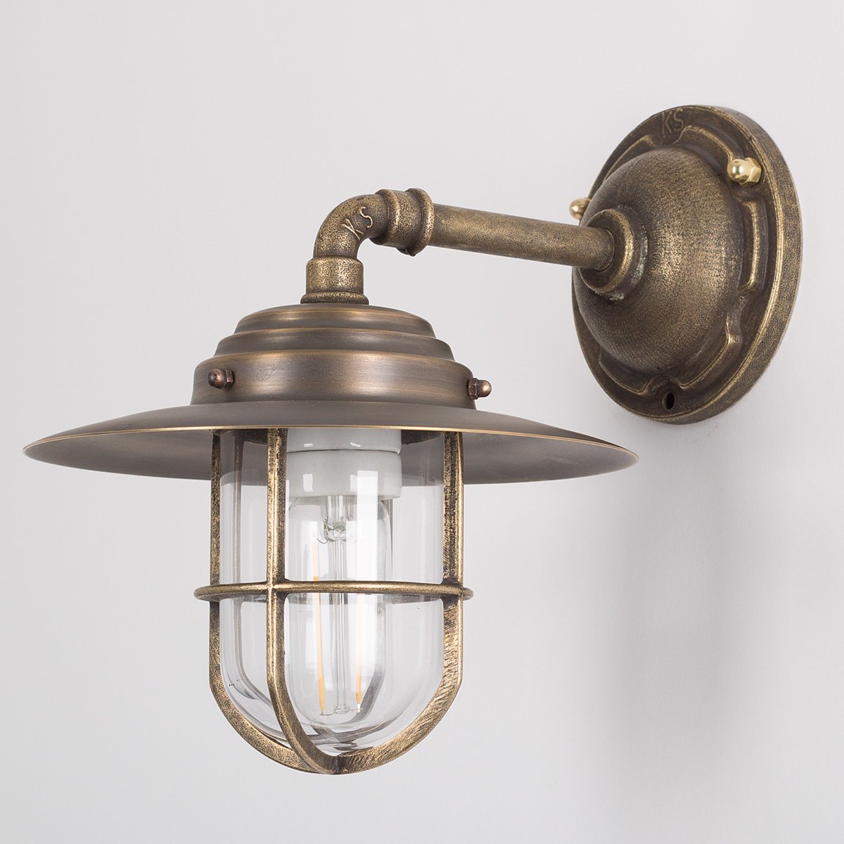 Buitenlamp Tour met bronzen wandarm, koperen kap, helder stolpglas en beschermend raster.